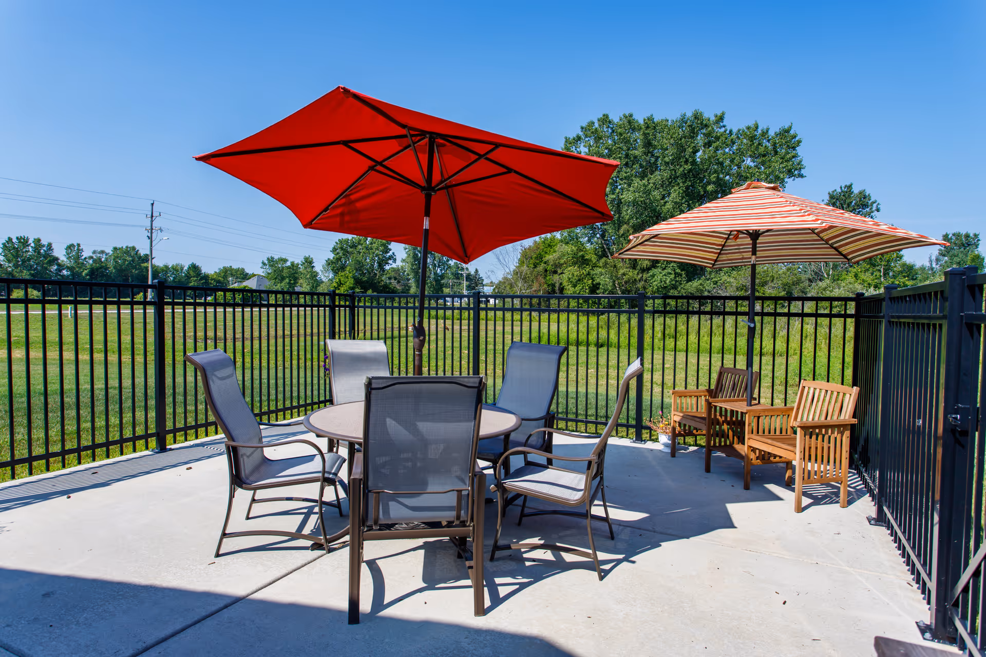 Fenced outdoor patio with a round table, several chairs, and red and striped umbrellas overlooking a grassy field.