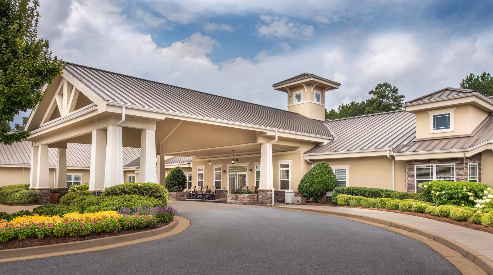 Front entrance of a low-rise building with a covered porte-cochère, landscaped flowerbeds, and a curved driveway.