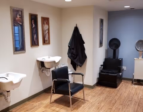 Interior of a senior living facility hair salon area with two white wash basins mounted on the wall, a black salon chair in front of one basin, a black hair drying chair with a hood dryer, and framed portraits on the walls. The floor is wooden and there are black salon capes hanging on the wall.