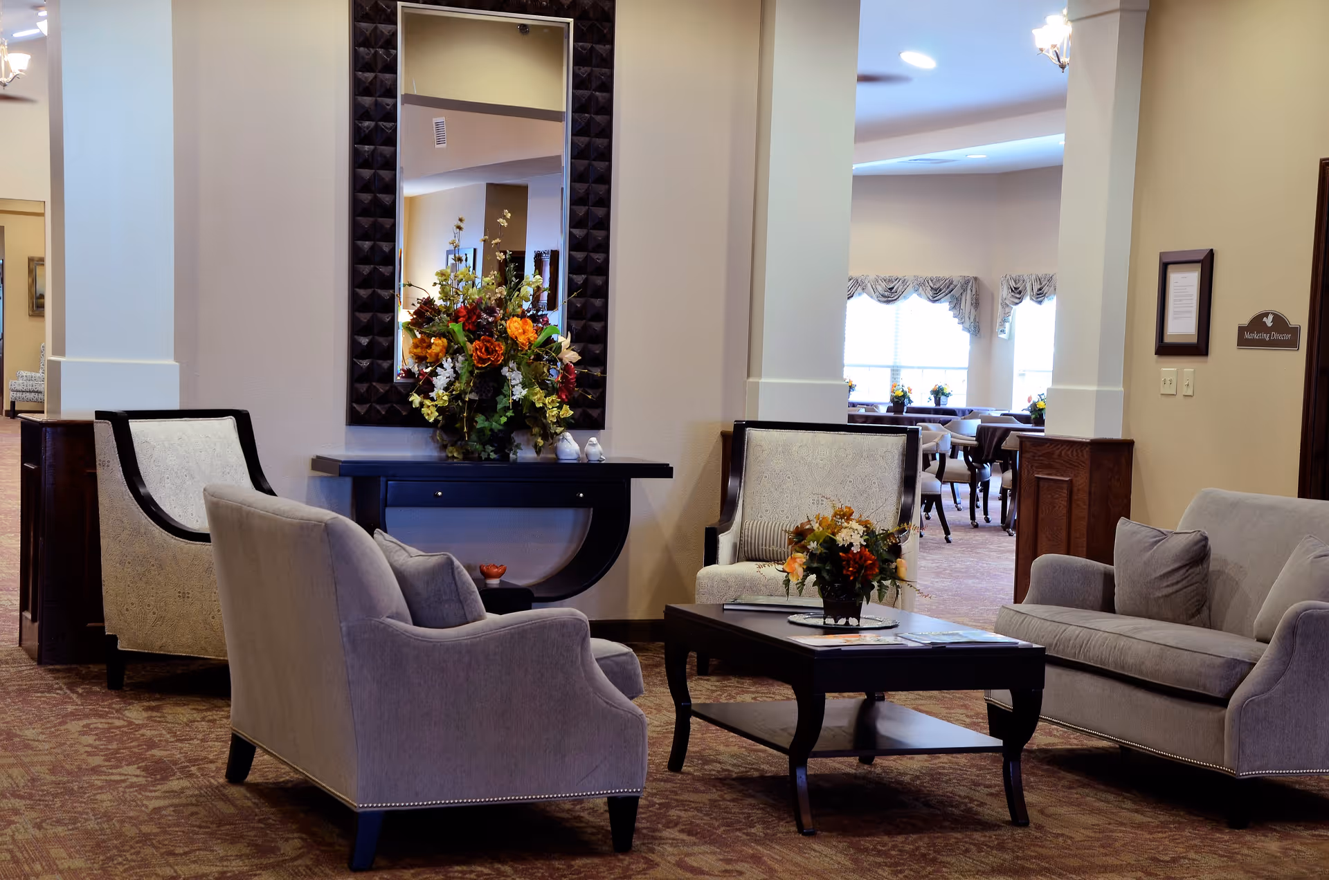 A cozy living room area in Culpepper Place featuring two armchairs and a sofa arranged around a dark wooden coffee table with a floral centerpiece. A console table with a large mirror and a flower arrangement is against the wall. In the background, a dining room with tables and chairs is visible through an open doorway.