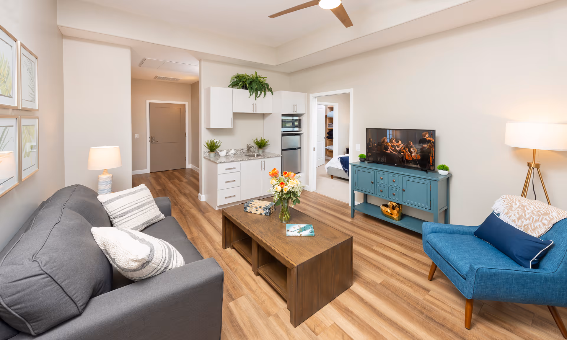 A cozy living room in an assisted living facility featuring a gray sofa with striped pillows, a wooden coffee table with a vase of flowers and books, a blue armchair with cushions, a blue TV stand with a flat-screen TV showing a string quartet, and a small kitchenette area with white cabinets and a mini fridge. The room has wooden flooring, neutral-colored walls, framed botanical prints, and a floor lamp.