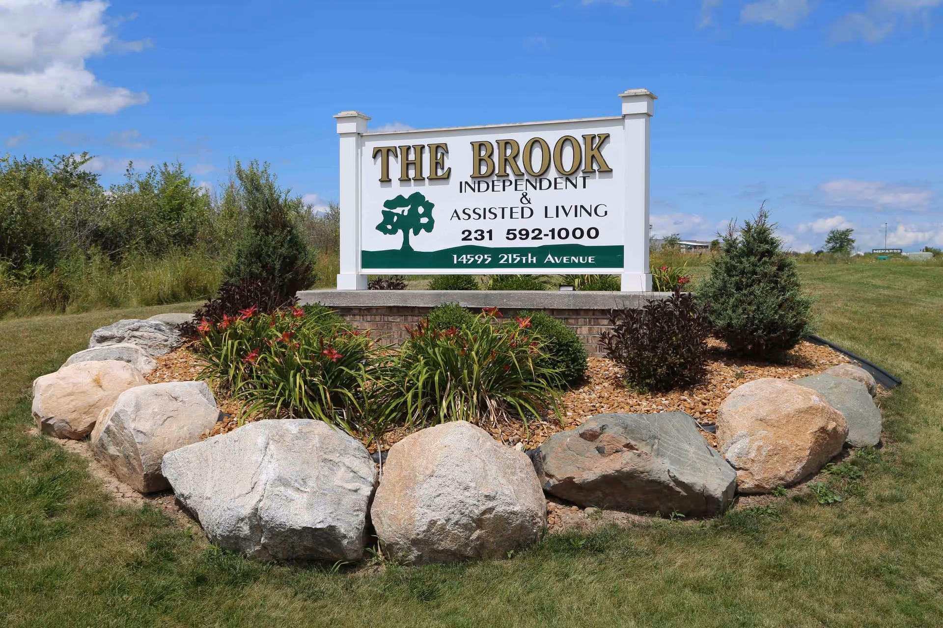 Outdoor landscaped area with large rocks and plants surrounding a white sign that reads 'THE BROOK INDEPENDENT & ASSISTED LIVING 231 592-1000 14595 215TH AVENUE' under a blue sky with some clouds.