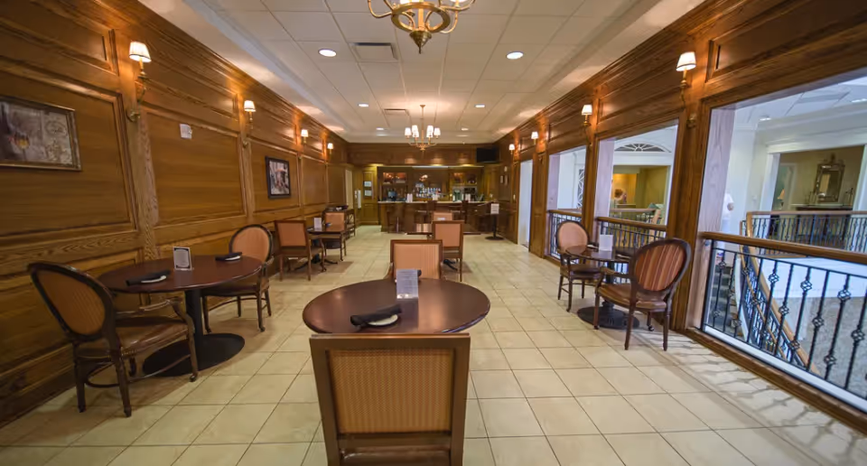 Interior view of a dining area with round wooden tables and upholstered chairs arranged neatly. The room features wood-paneled walls with framed artwork and wall sconces, tile flooring, and large windows on the right side overlooking an interior balcony. At the far end, there is a bar area with shelves and a chandelier hanging from the ceiling.