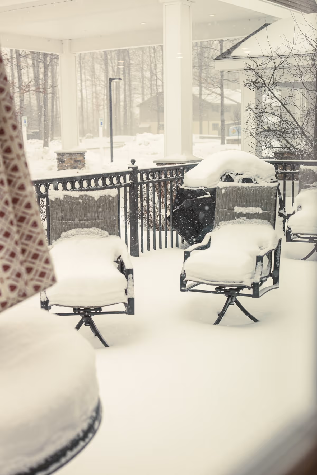 Snow-covered outdoor patio with two chairs and a grill, surrounded by a black metal fence and white pillars, with snow falling and trees in the background.