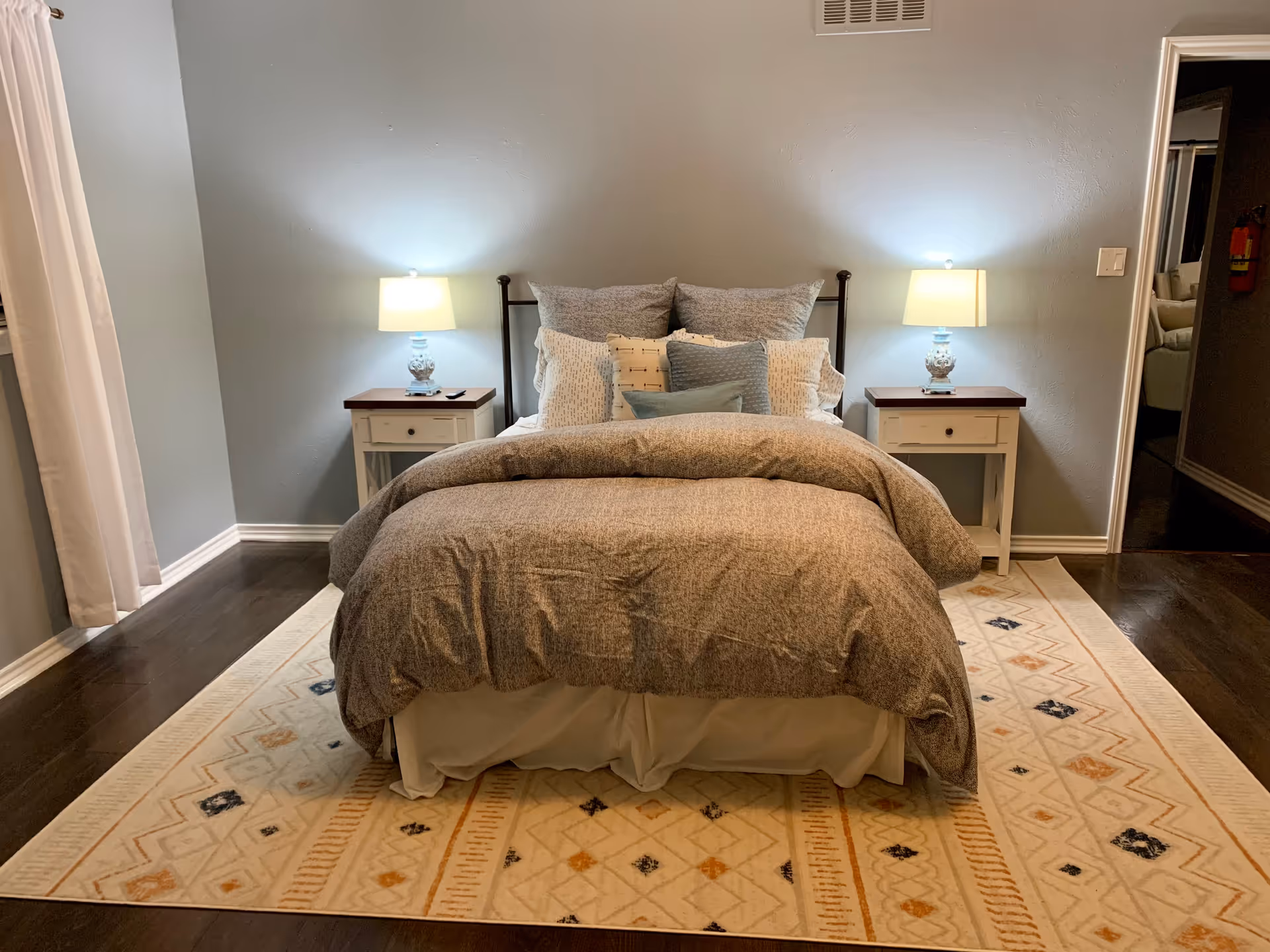 A neatly made bed with multiple pillows and a gray comforter in a bedroom. The bed is flanked by two white nightstands, each with a lamp emitting warm light. The room has light blue walls, dark hardwood floors, and a patterned area rug under the bed. A white curtain is partially visible on the left side of the image.
