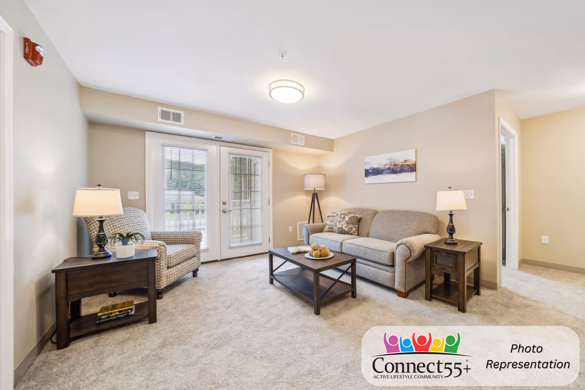 A bright and cozy living room in a senior living community featuring a beige sofa with patterned cushions, a patterned armchair, two wooden side tables each with a lamp, a wooden coffee table with decorative items, and glass double doors with blinds leading outside. The room has beige walls and carpeted flooring.