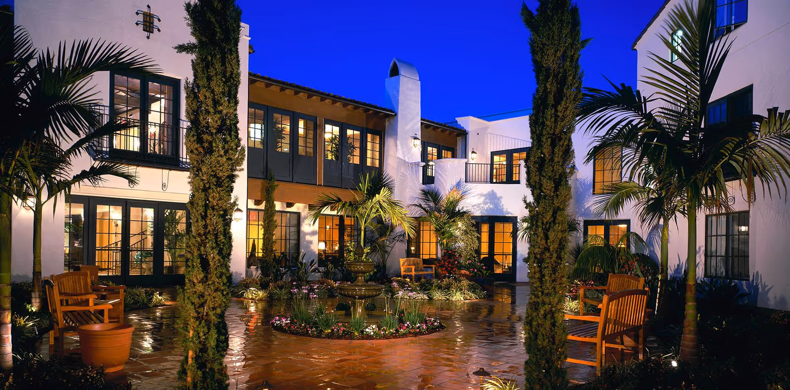 Evening view of a courtyard at the Maravilla facility featuring a central fountain surrounded by flowers and plants, wooden benches, tall trees, and a white two-story building with large windows illuminated from inside.