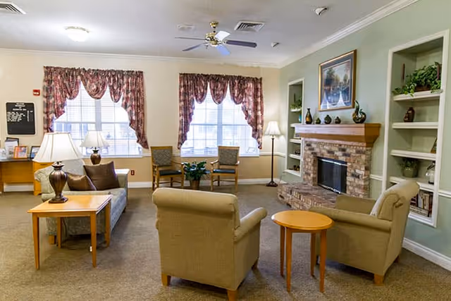 A cozy living room with two beige armchairs and a sofa arranged around a brick fireplace. The room has built-in shelves with decorative items, two large windows with floral curtains, a floor lamp, and a table lamp on a side table next to the sofa.