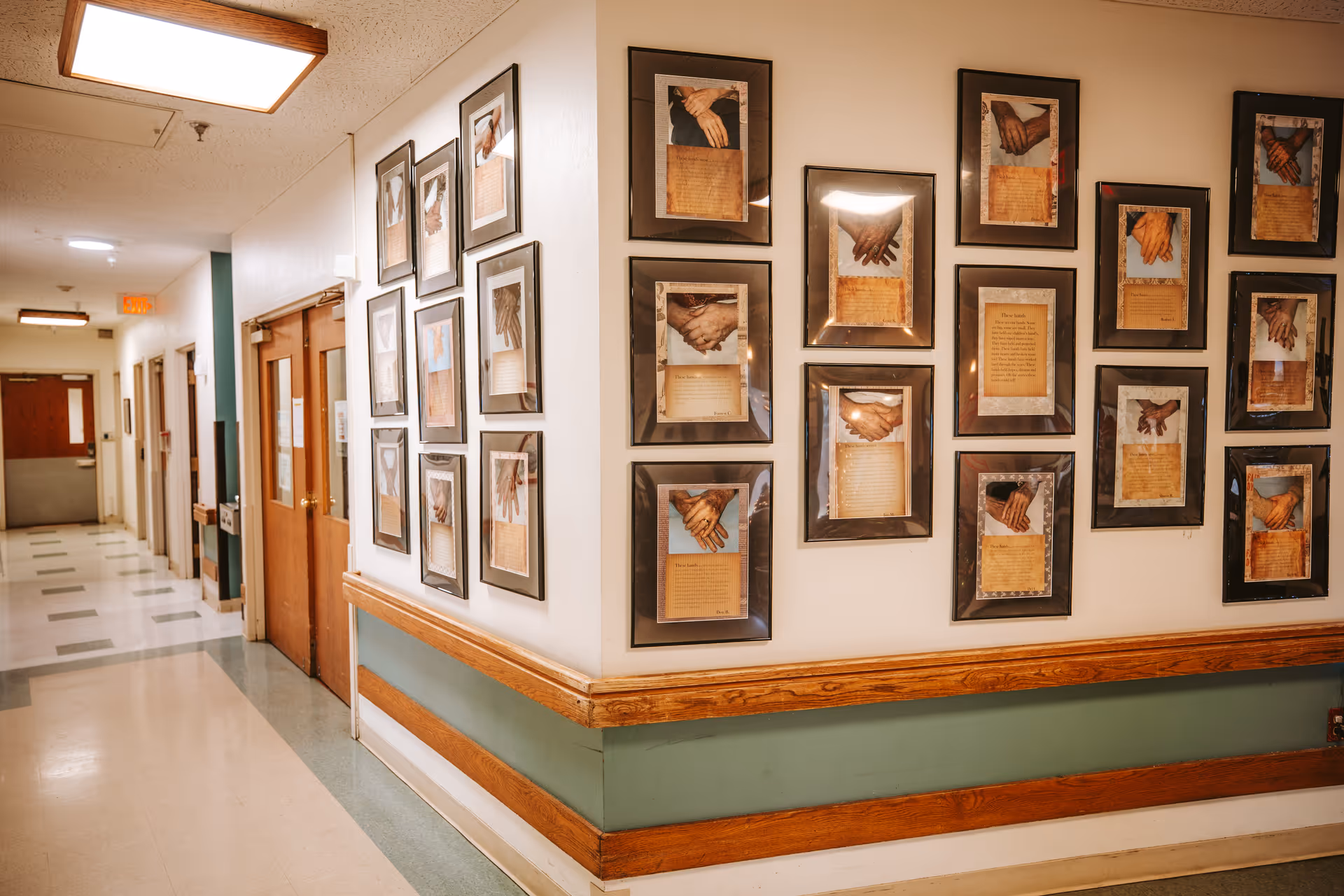 A hallway in Cooney Healthcare and Rehabilitation with framed pictures of hands holding each other and accompanying text on the walls. The hallway has a green and beige color scheme with wooden trim and several doors along the corridor.