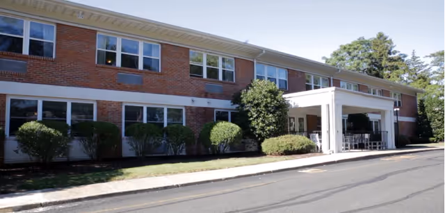 Exterior view of a two-story brick building with multiple windows and a covered entrance. There are bushes and small trees planted along the front of the building, and a paved driveway or road runs alongside it.