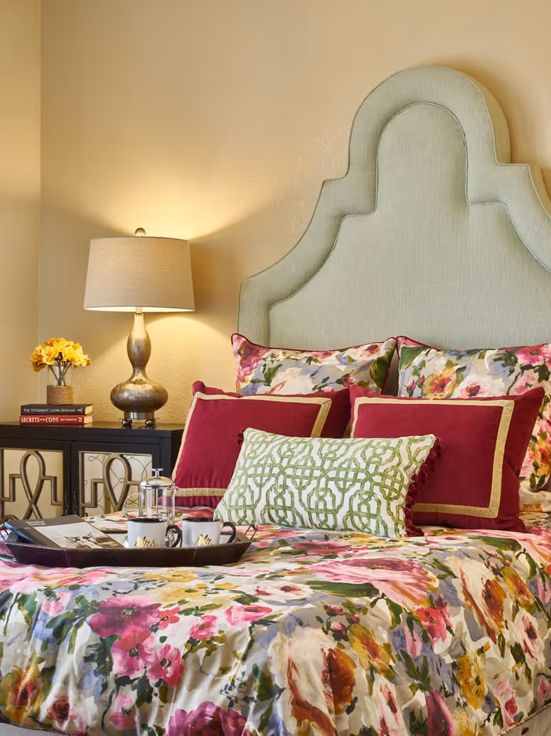 A cozy bedroom featuring a bed with a light green upholstered headboard, floral bedspread, and multiple decorative pillows in red and green patterns. A bedside table with a lamp, a small vase of yellow flowers, and two books is visible next to the bed. A tray with two coffee mugs and a French press sits on the bed.