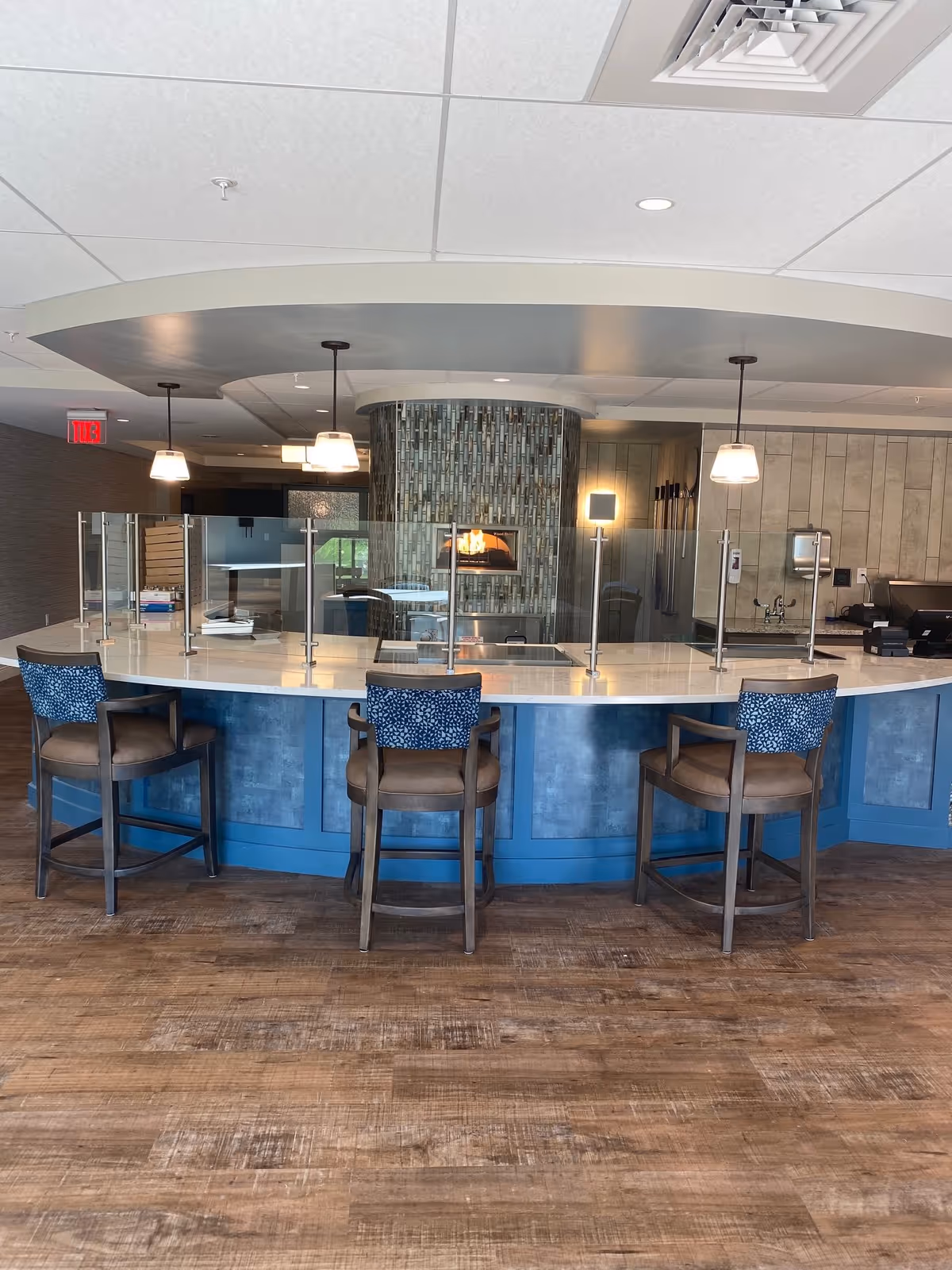 A modern curved counter with three high chairs featuring blue patterned cushions. The counter has a glass partition on top. Behind the counter is a fireplace with a tiled surround and a wood-fired oven. The floor is wooden, and there are pendant lights hanging from the ceiling.