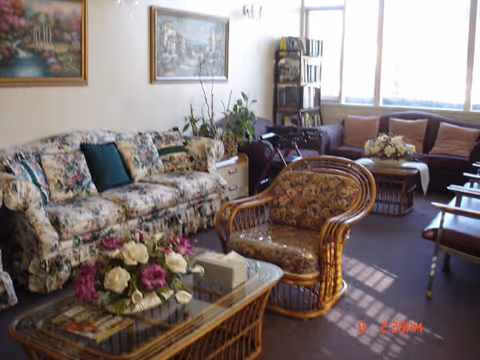 A cozy living room with floral patterned sofa and armchair, a glass-top wicker coffee table with a flower arrangement, additional seating with a dark sofa and chairs, large windows letting in natural light, and framed paintings on the wall.