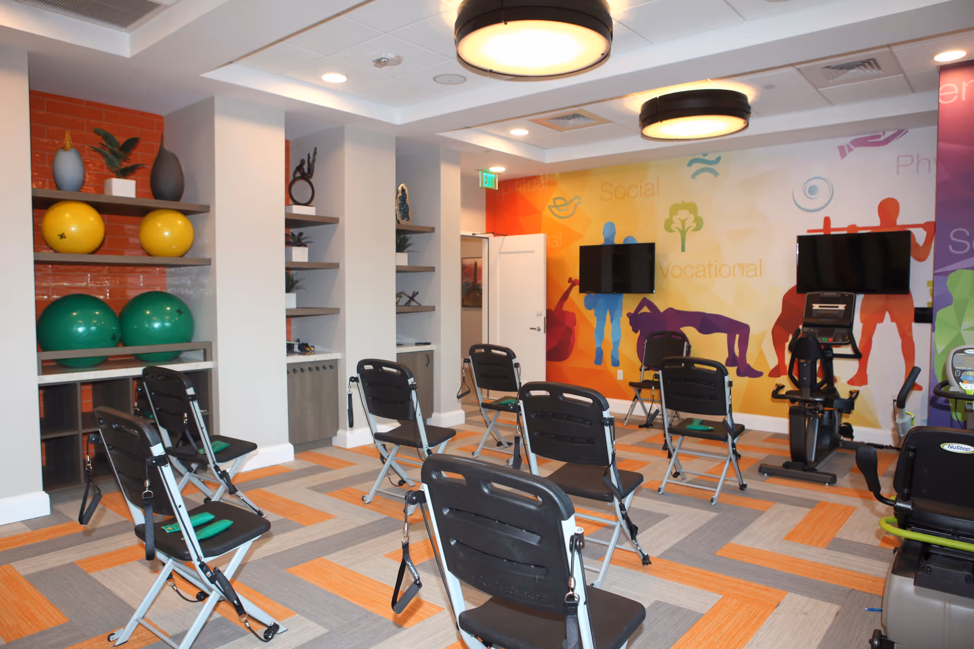 A fitness room with several black folding exercise chairs arranged in rows, each equipped with resistance bands. The room has colorful wall art featuring silhouettes of people exercising and words like 'Social' and 'Vocational'. There are two flat-screen TVs mounted on the wall and exercise equipment including a recumbent bike. Shelves on the left hold large yellow and green exercise balls and decorative items. The floor has a patterned carpet with orange, gray, and beige tones.