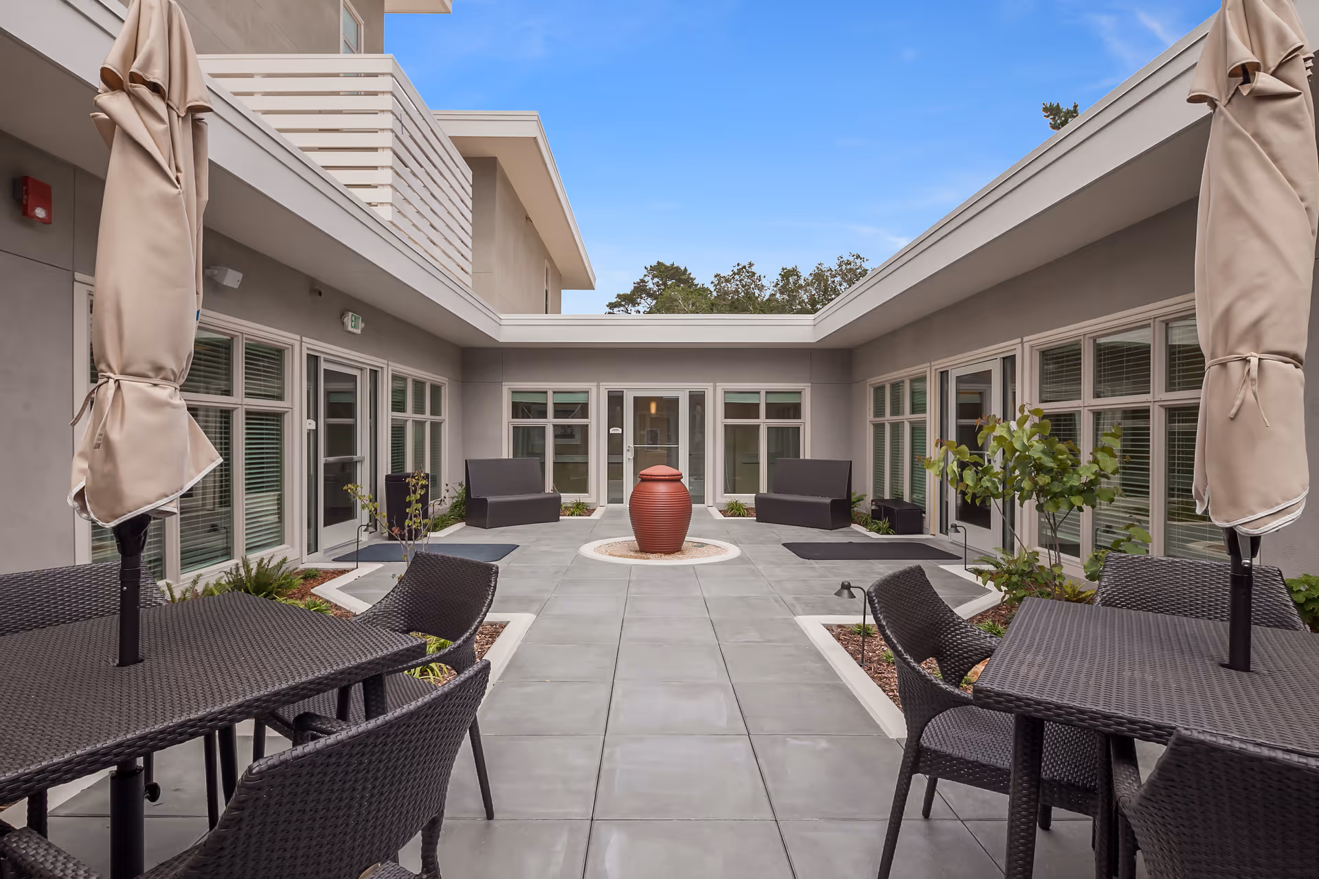 Outdoor courtyard area with tiled flooring, two black wicker tables with umbrellas and chairs, two black benches, potted plants, and a large red decorative urn in the center. The courtyard is surrounded by a building with multiple windows and doors under a clear blue sky.