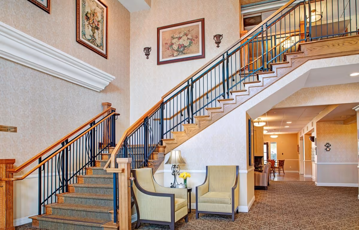 Interior view of a senior living facility featuring a carpeted staircase with wooden handrails and black metal balusters. Two beige armchairs are placed beside a small table with a lamp and a vase of yellow flowers. The walls are decorated with framed floral artwork and sconces. The hallway extends into the background with additional seating and lighting fixtures.