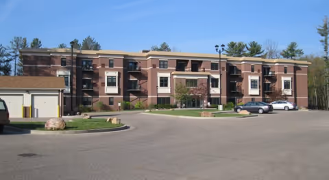 Exterior view of a three-story brick assisted living facility building with balconies, surrounded by trees and a parking lot with several cars parked.