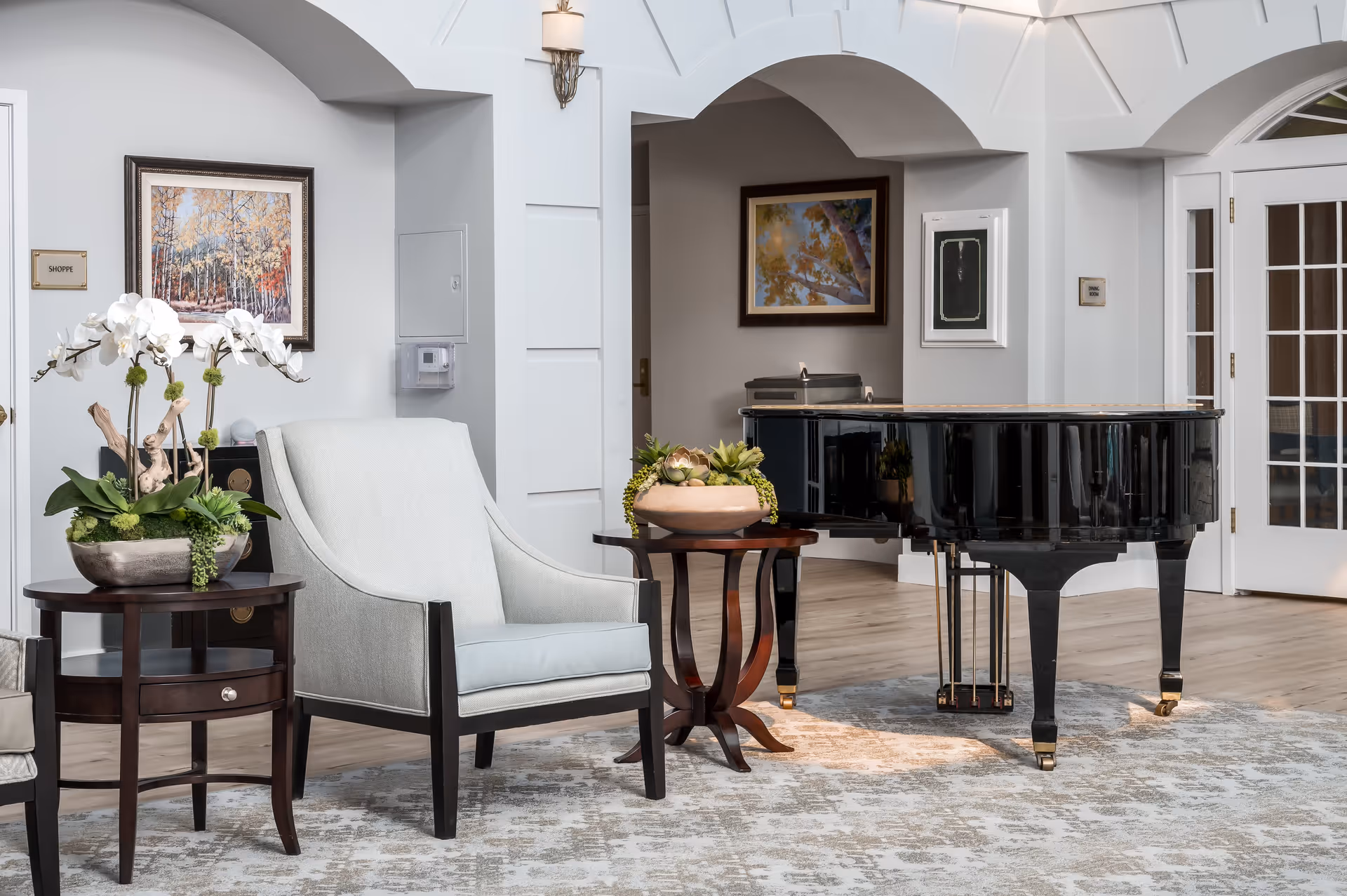 A bright and elegant interior room featuring a black grand piano on a patterned rug, a light gray upholstered armchair, and two dark wooden side tables with decorative plants. The walls are white with framed artwork and sconces, and there are white double doors with glass panes on the right side.