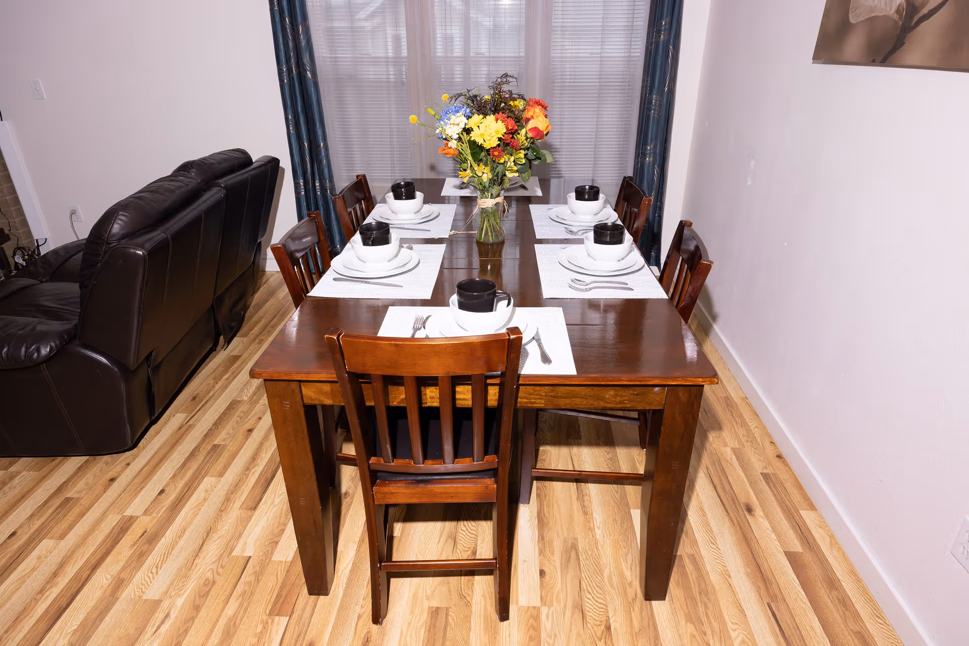 A wooden dining table set for six with white place settings and a floral centerpiece in a dining area next to a leather couch.