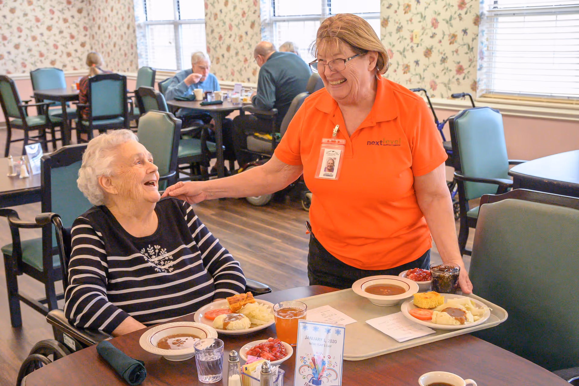 An elderly woman in a wheelchair is smiling and interacting with a caregiver wearing an orange shirt and name badge in a dining room. The caregiver is holding a tray with plates of food and drinks. Other elderly people are seated at tables in the background, eating and conversing. The room has floral wallpaper, large windows, and green cushioned chairs.