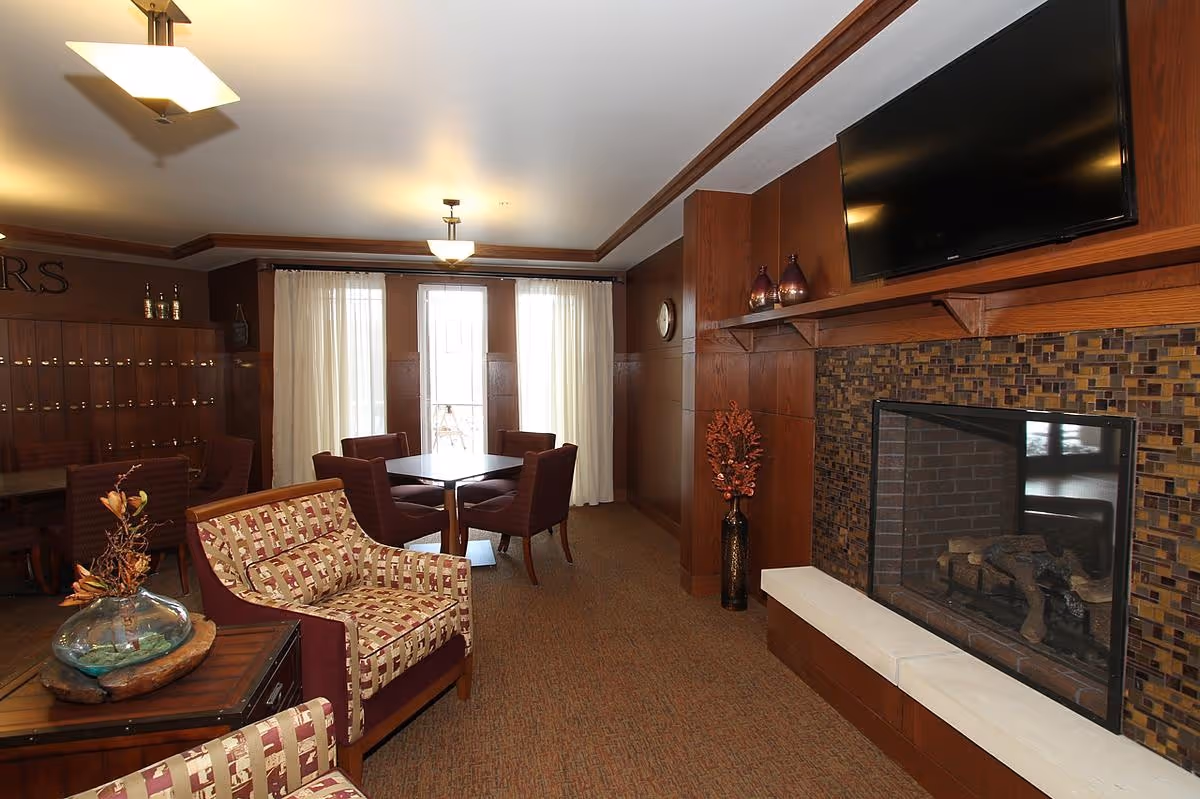 Cozy common room with patterned armchairs, a dining table, fireplace with a tiled surround, and a wall-mounted TV.