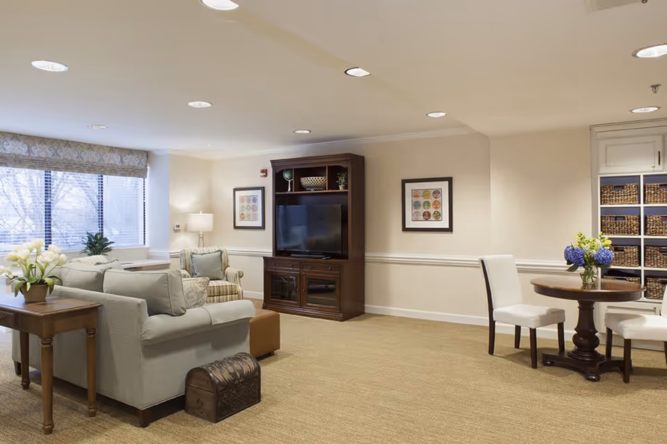 A cozy living room area in a senior living facility featuring a light gray sofa with cushions, a wooden side table with a flower arrangement, a striped armchair, a wooden entertainment center with a flat-screen TV, framed artwork on the walls, a round wooden table with two white chairs, and shelves with woven baskets. Large windows with patterned valances allow natural light to fill the room.