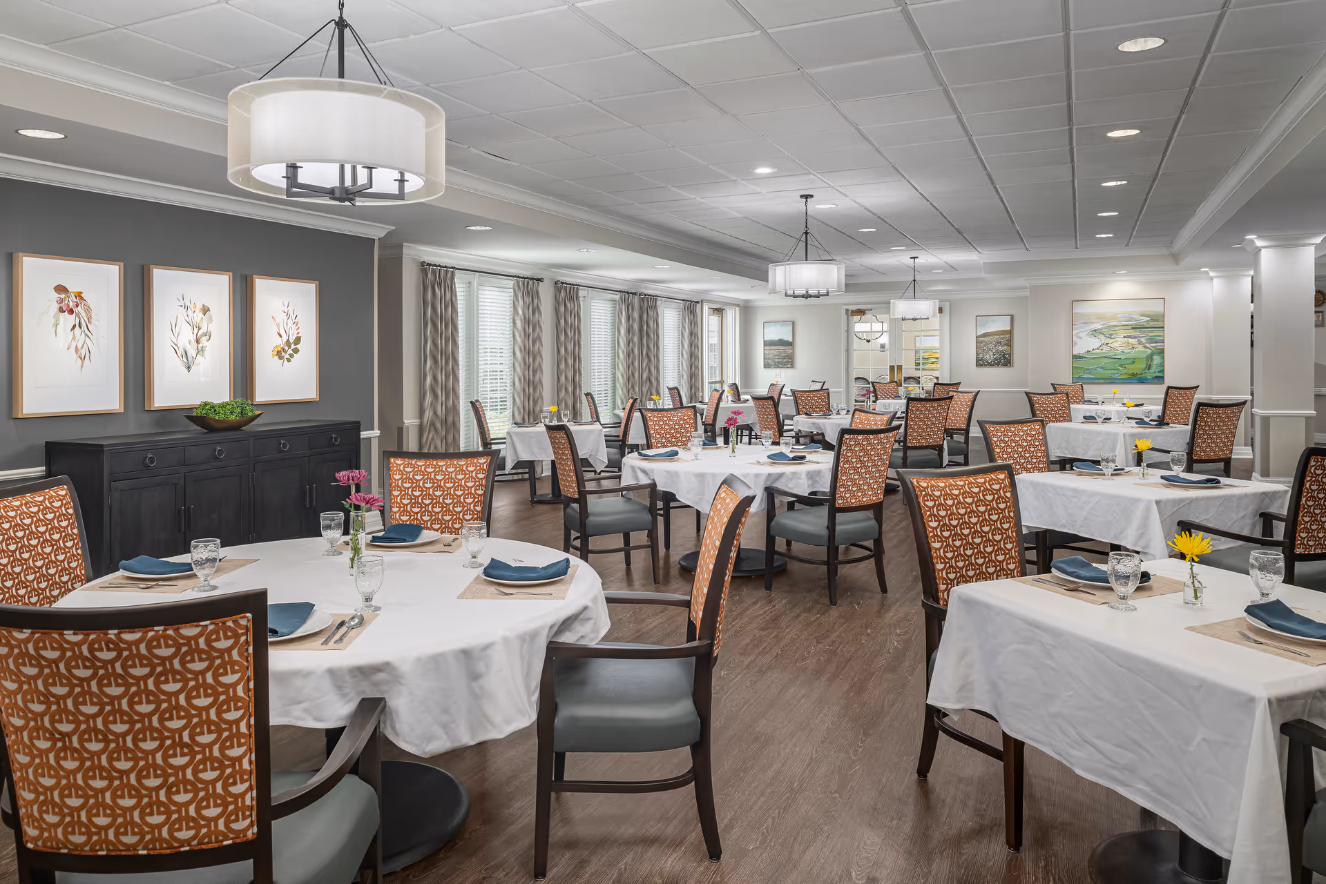 A spacious dining room with multiple round and square tables covered with white tablecloths. Each table is set with blue napkins, glassware, and small flower vases. The chairs have patterned orange backs and gray seats. The room has large windows with patterned curtains, framed botanical artwork on the walls, and modern ceiling lights.