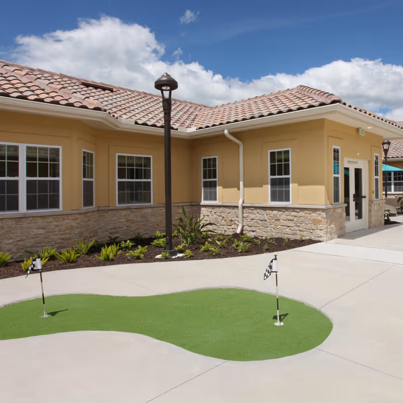 Outdoor courtyard area of a senior living facility with a small putting green, two golf flags, beige building with stone accents, multiple windows, a lamp post, and a partly cloudy sky.