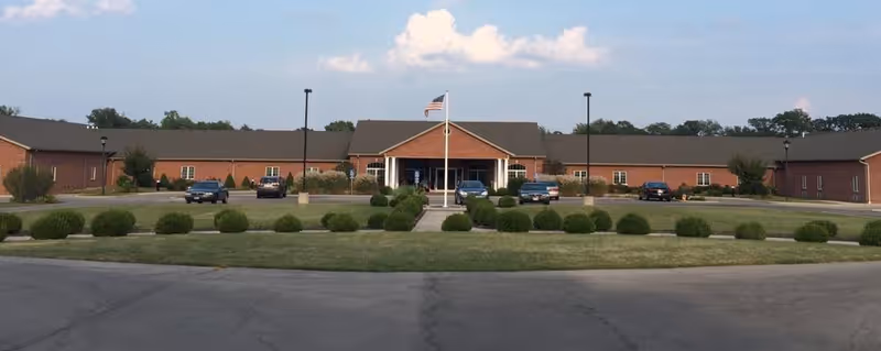 Wide front view of a single-story brick assisted living building with a central entrance, flagpole, and circular driveway with parked cars.