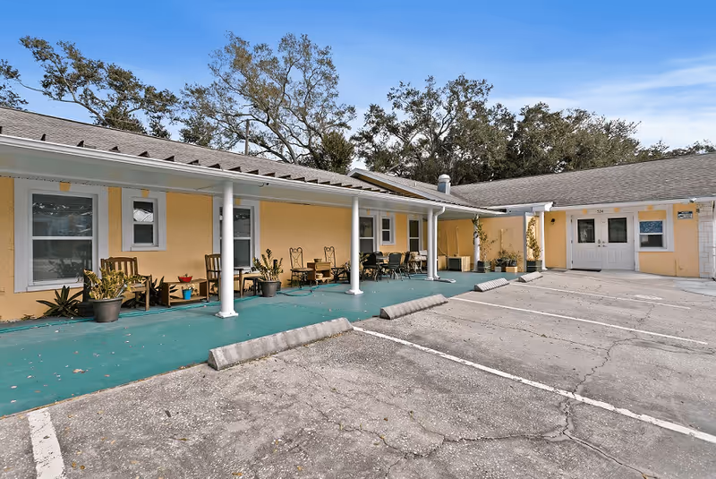 Exterior view of a single-story building with a covered porch area featuring several chairs, tables, and potted plants. The building is painted yellow with white trim and has a parking area in front with marked parking spaces. Trees are visible in the background under a clear blue sky.