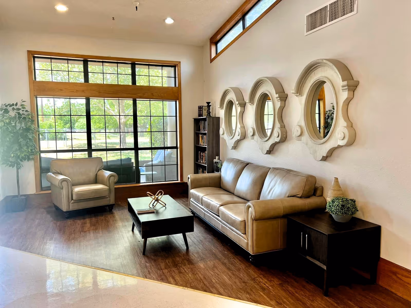 A cozy living room area with a large window showing greenery outside. The room features a beige leather sofa, a matching armchair, a dark wooden coffee table with decorative items, a small bookshelf, and three ornate round mirrors on the wall above the sofa. There is also a small side table with a plant and a decorative vase.