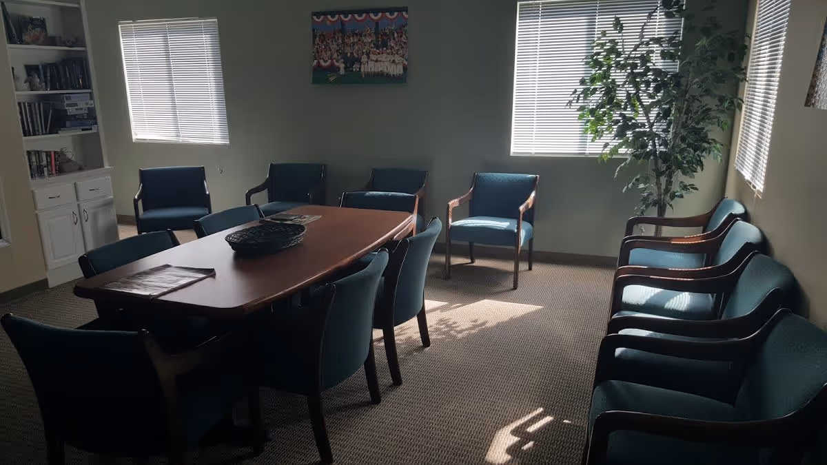 A well-lit room with a long wooden table surrounded by blue upholstered chairs. Additional blue chairs are arranged along the walls. Two windows with blinds allow sunlight to enter, casting shadows on the carpeted floor. A tall potted plant is placed near one window, and a bookshelf with books and decorative items is visible in the corner.