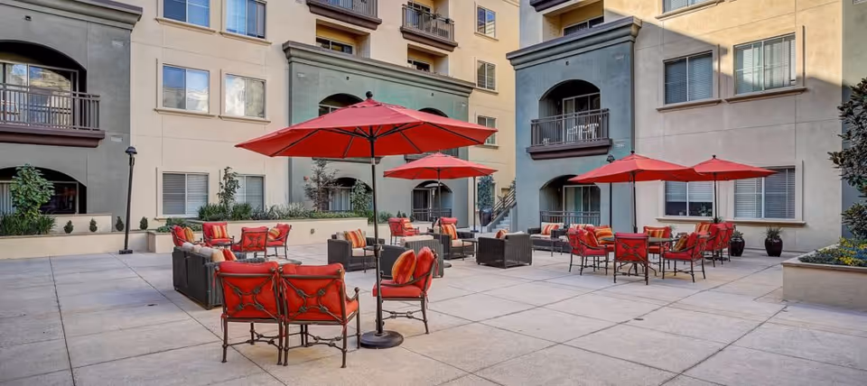 Outdoor courtyard with red umbrellas, cushioned chairs and tables surrounded by a multi-story residential building.