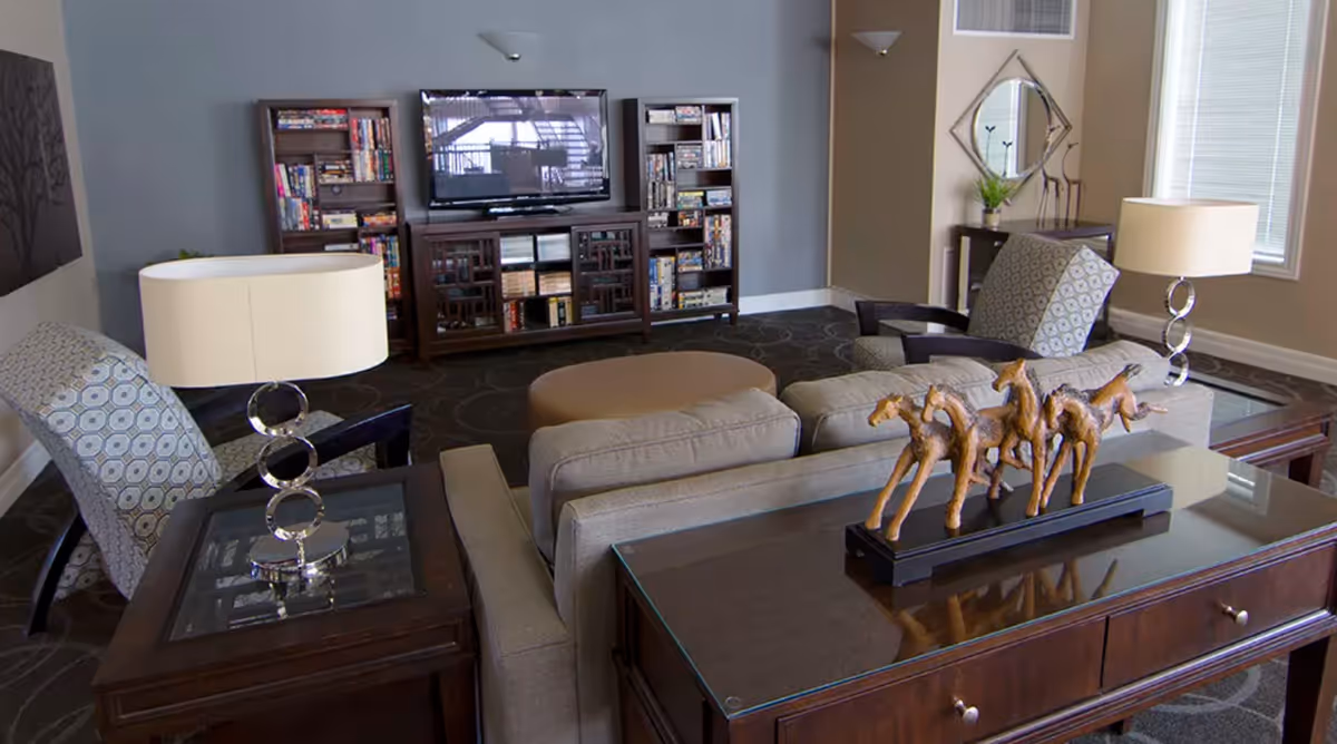 Communal living room with sofas, armchairs, side tables and lamps facing a TV and bookshelf, with decorative horse sculptures on a console table.