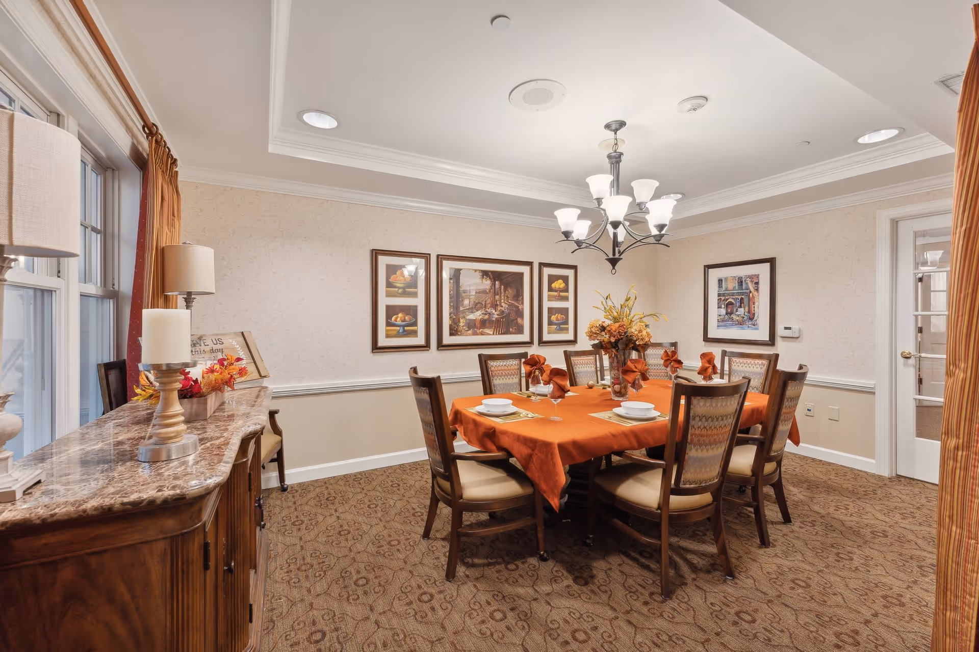 A dining room with a rectangular table covered with an orange tablecloth, set with white plates, bowls, and orange napkins. Eight wooden chairs with cushioned seats surround the table. A chandelier with multiple lights hangs above the table. The room has beige walls adorned with framed artwork and a patterned carpet. A wooden sideboard with a marble top holds decorative items including a lamp and candles. Large windows with orange curtains allow natural light into the room.