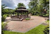 A paved outdoor courtyard area featuring a wooden gazebo surrounded by trees and greenery under a partly cloudy sky.