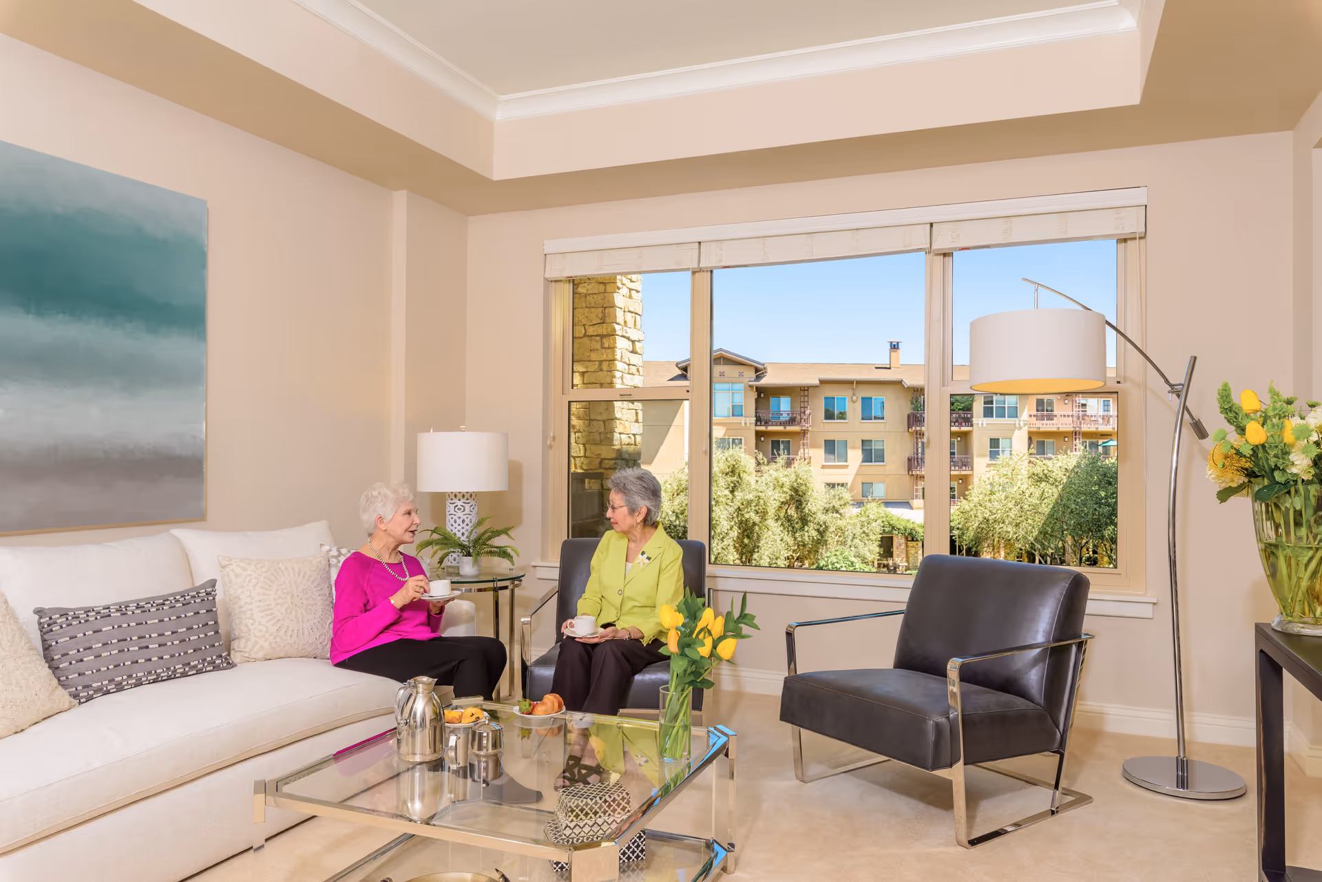 Two elderly women sitting and having tea in a bright living room with large windows showing an outside view of a building and greenery. The room has a white couch with pillows, a glass coffee table with a tea set and pastries, a modern floor lamp, and a vase with yellow tulips.