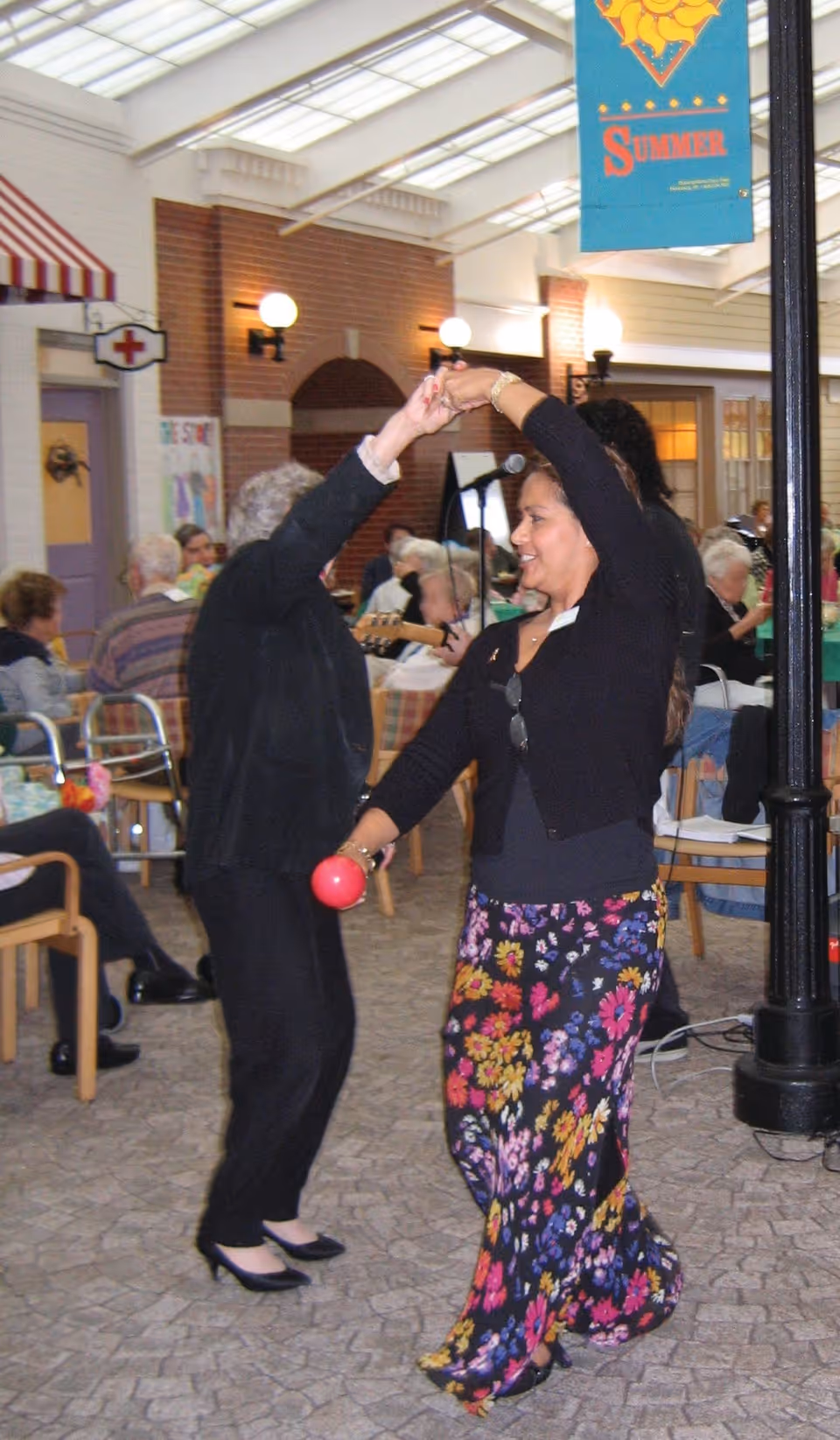 Two women dancing indoors in a lively room filled with seated elderly people. One woman is wearing a black outfit and the other is wearing a black top with a colorful floral skirt, holding a red maraca. The room has a cozy atmosphere with brick walls, warm lighting, and a banner that says 'Summer' hanging from the ceiling.