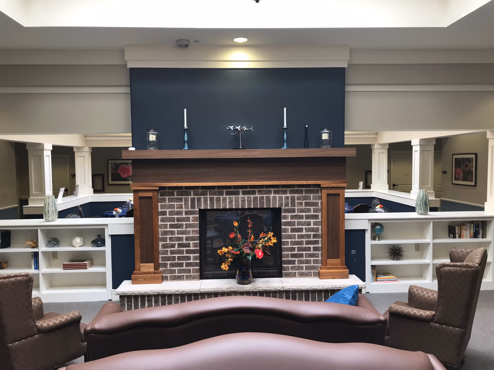 A cozy living room area in a senior living facility featuring a brick fireplace with a wooden mantle. The mantle is decorated with two blue candle holders, a small sculpture, and glass jars. In front of the fireplace is a vase with colorful flowers. Surrounding the fireplace are built-in white shelves with decorative items and books. There are two brown armchairs and a brown sofa facing the fireplace. The walls are painted in neutral tones with a dark blue accent behind the fireplace.