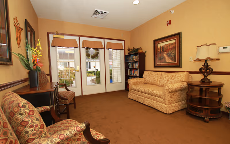 A cozy living room area with a patterned beige sofa, two patterned armchairs, a wooden side table with a lamp, a bookshelf filled with books, and a framed painting on the wall. The room has beige walls, brown carpet, and glass doors with valances leading outside.