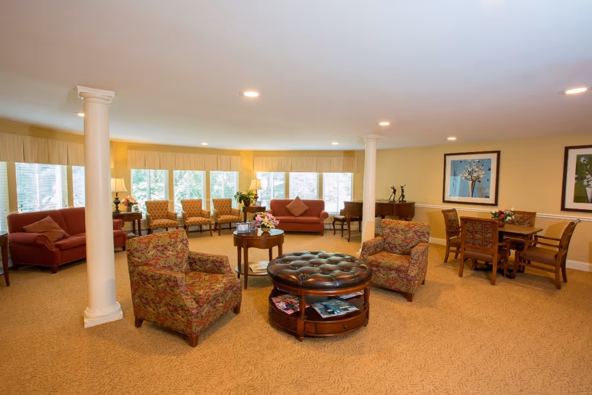 A spacious senior living common area with beige carpeting and yellow walls. The room features multiple seating options including two patterned armchairs, two red sofas, and four wooden chairs with patterned cushions. There is a round leather ottoman with magazines underneath in the center, a small round wooden table with flowers, and a wooden dining table with four chairs. Large windows with blinds and valances let in natural light. Decorative columns, framed artwork, table lamps, and a piano add to the cozy atmosphere.