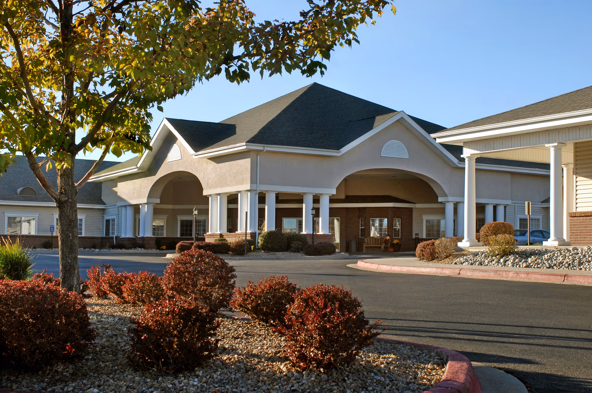 Exterior view of the Life Care Center of Westminster building entrance with a covered driveway, white columns, and landscaped bushes and trees in the foreground under a clear blue sky.