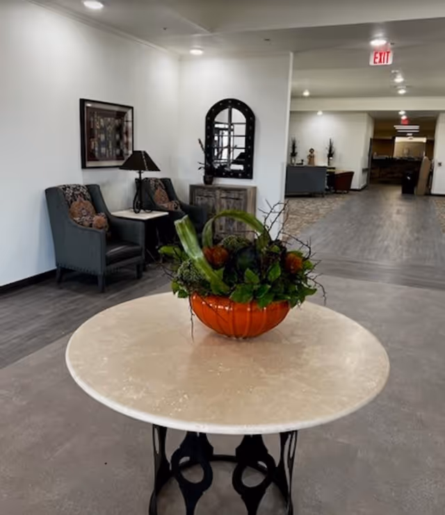 Round table with an orange decorative bowl of plants in a lobby/common area with seating and a hallway behind it.