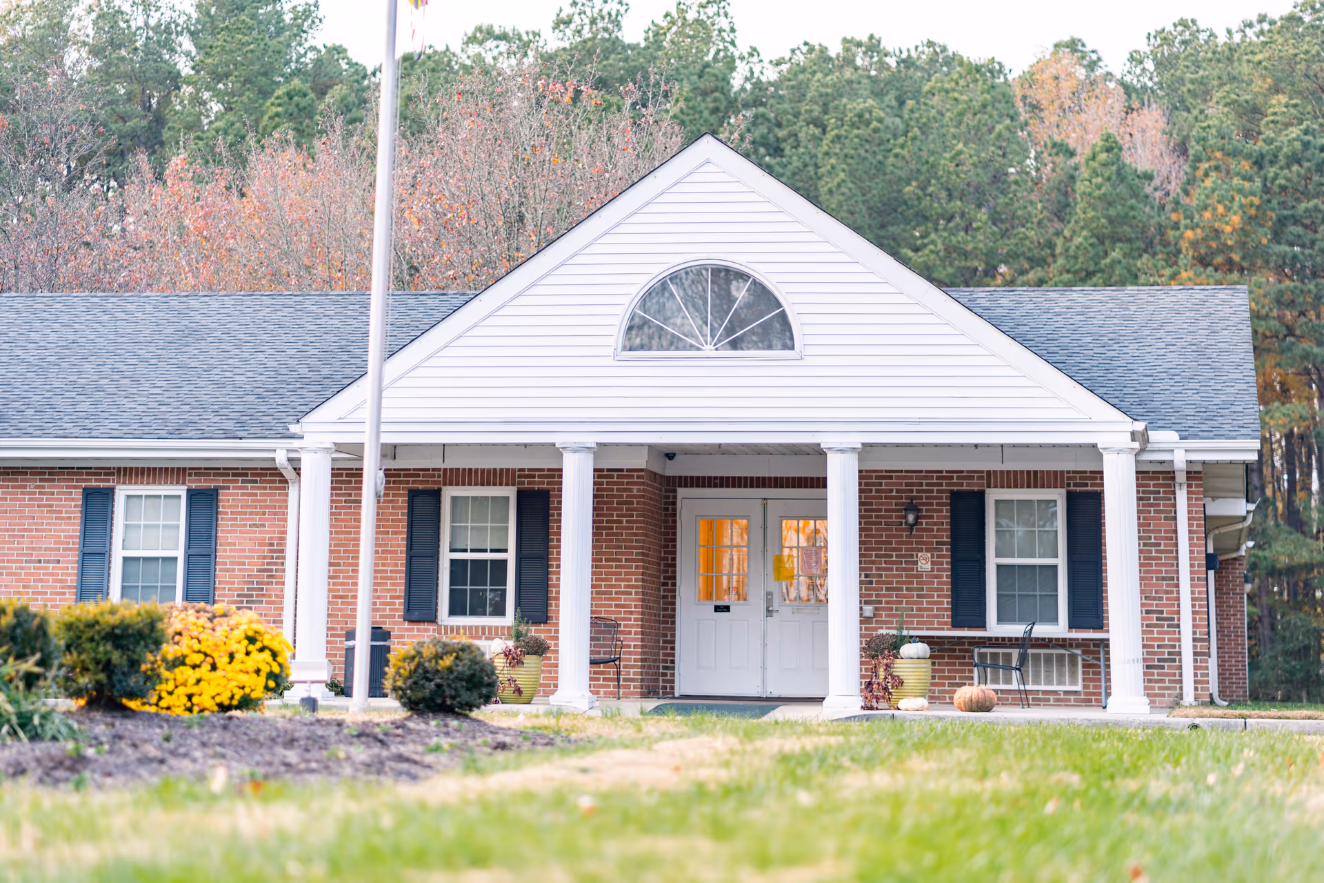 Front exterior view of a single-story brick building with white columns and a triangular pediment above the entrance. The building has white double doors with glass panels, black shutters on the windows, and is surrounded by green grass, shrubs, and yellow flowers. Trees with autumn foliage are visible in the background.