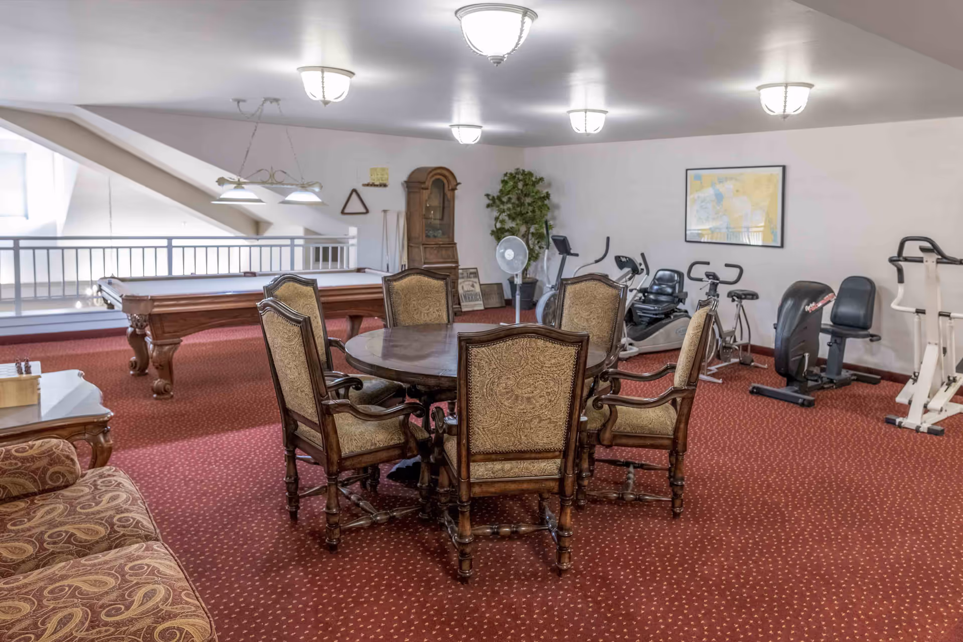 A carpeted common room with a round table and six upholstered chairs, a pool table, and various exercise machines.