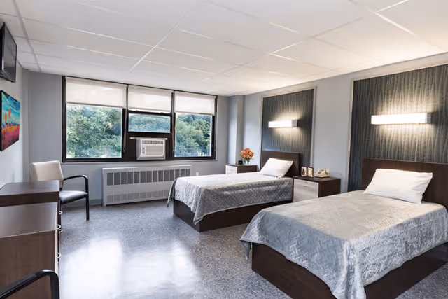 A bright and clean bedroom in a senior living facility with two single beds, each with a gray bedspread and a pillow. The room has large windows with a view of green trees outside, an air conditioning unit below the windows, a chair, a dresser, and two nightstands with a telephone, framed photos, and a vase of flowers. The walls are painted light blue with dark accent panels behind the beds, each featuring a modern wall light.