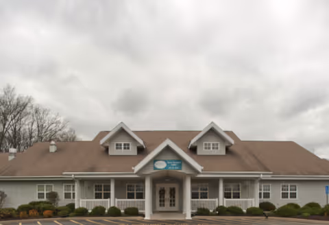 Front exterior view of a single-story building with a brown roof and white siding, featuring a covered entrance with two columns and a sign above the door. The building is surrounded by landscaped bushes and a parking lot under a cloudy sky.
