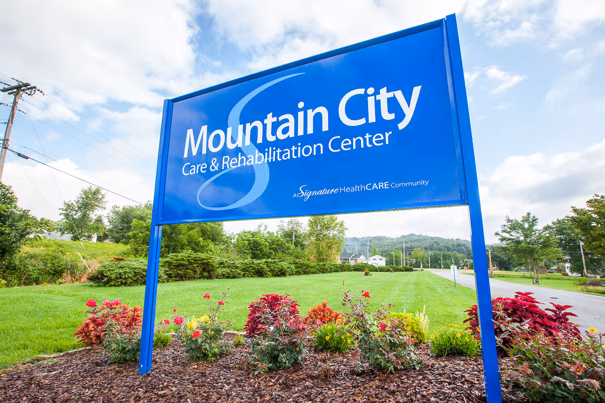 Blue roadside sign reading 'Mountain City Care & Rehabilitation Center' standing in a landscaped lawn with flowers and trees.