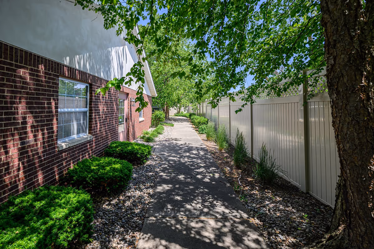 A paved walkway beside a brick and white building with windows and a door. The walkway is lined with green bushes and small rocks on one side and a white fence with plants on the other. Trees with green leaves provide shade over the path under a clear blue sky.