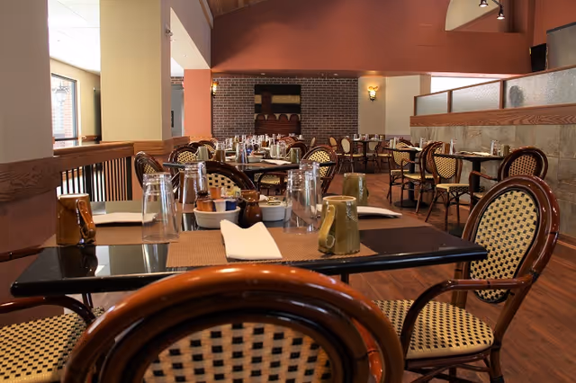 Interior view of a dining room with multiple tables and chairs arranged neatly. Each table is set with placemats, napkins, glasses, and mugs. The room has warm lighting, wooden flooring, and a decorative brick wall in the background.